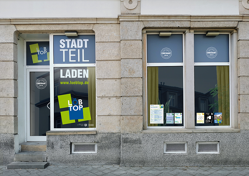 Fenster des Stadtteilladens des Löbtop e.V.s von außen. Links ist das Logo und groß "STADTTEIL LADEN" aufgeklebt. Rechts sind kleine Veranstaltungsplakate an der Schreibe.